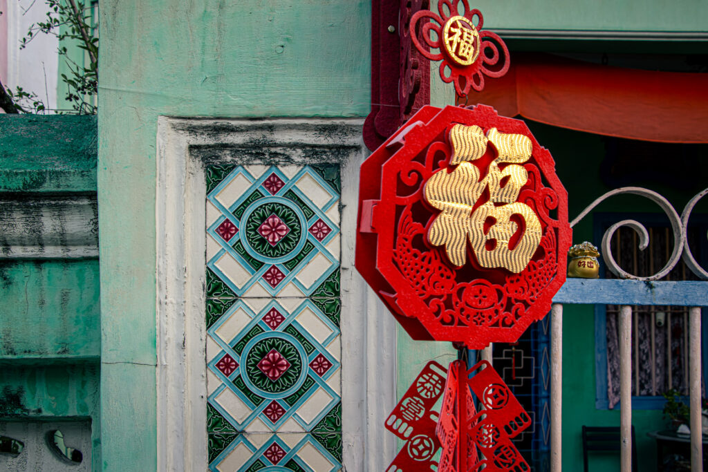 A close-up of a bright red Chinese New Year ornament with a gold 福 (fú) character catching the morning sun, hanging against a mint green wall with geometric encaustic tiles.
