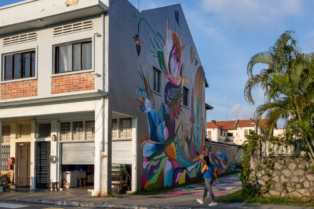 Large abstract floral mural in blues, pinks, and greens covering the side of a building, with a woman walking past below.