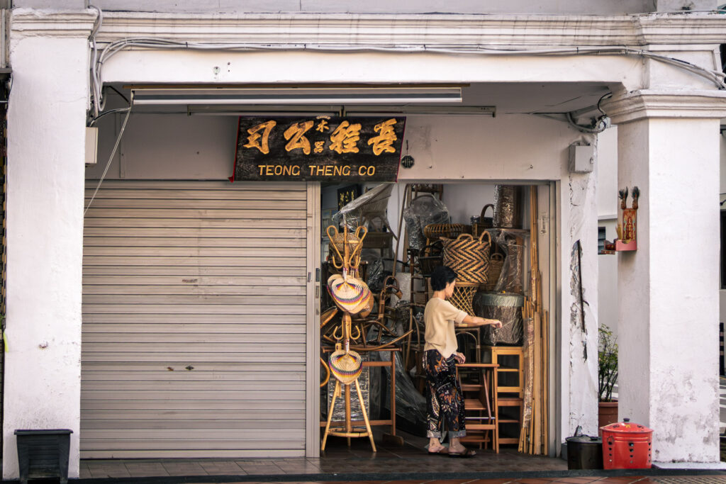 Open-fronted rattan and bamboo furniture shop with woven baskets and furniture on display, a woman arranging stock inside.