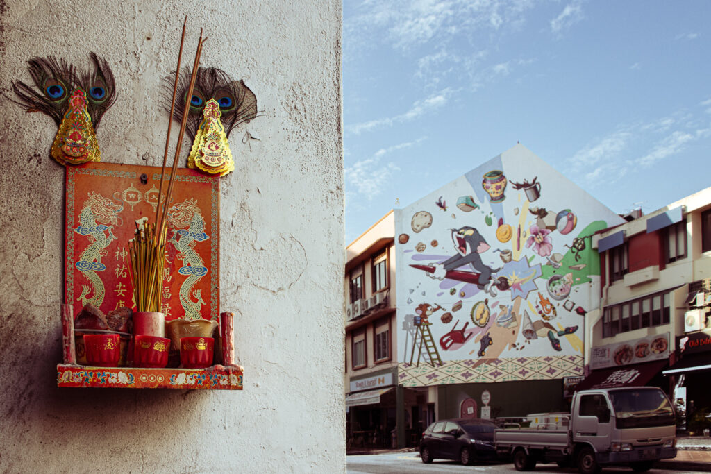 A wall-mounted red incense altar decorated with peacock feathers, across the street a colourful Tom and Jerry mural on a building facade.
