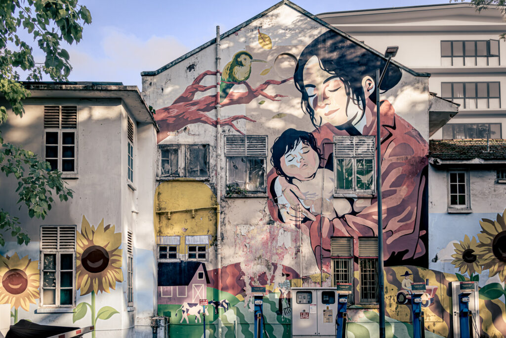 Weathered mural of a mother cradling an infant, painted on the peeling facade of a disused building.