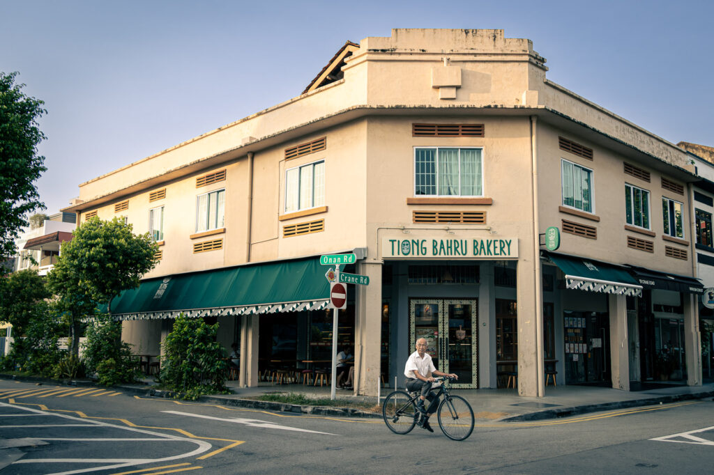 Cream corner building with Tiong Bahru Bakery signage; an elderly man cycles past on the street corner.