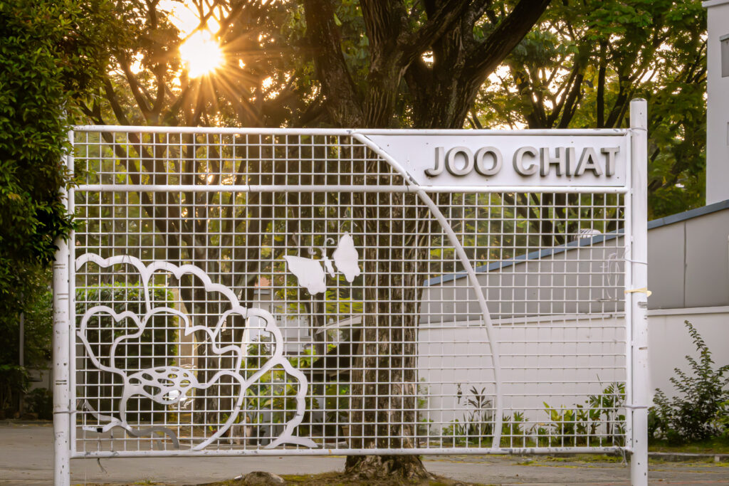 White metal gateway sign reading 'Joo Chiat' with a cut-out floral and butterfly design, backlit by the rising sun through trees.