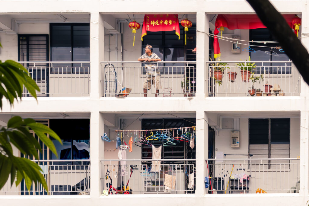 Elderly man on HDB flat balcony beneath red Chinese New Year banners, neighbouring balconies strung with laundry and potted plants, Singapore, from Kamsin Kaneko's LRPS panel