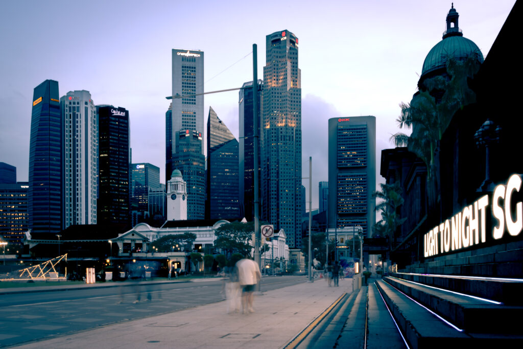 Singapore skyline at dusk with illuminated skyscrapers, National Gallery dome, and blurred pedestrians beneath a "Light to Night" light installation sign. From Kamsin Kaneko's LRPS panel