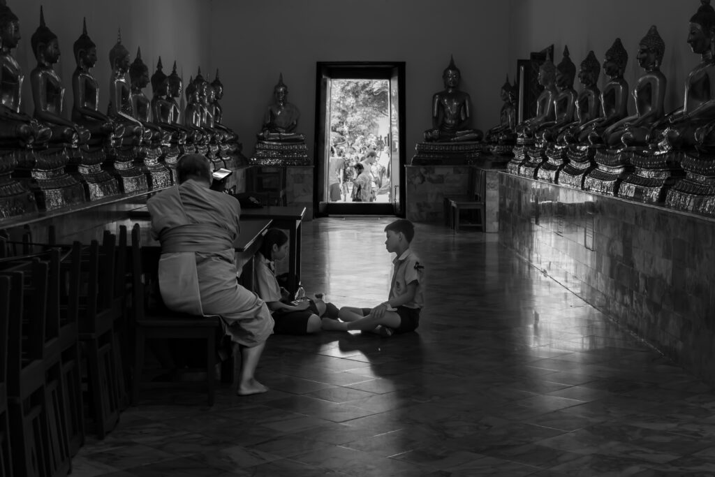 Monk and two children seated on the floor of a Buddhist temple hall lined with rows of seated Buddha statues, black and white